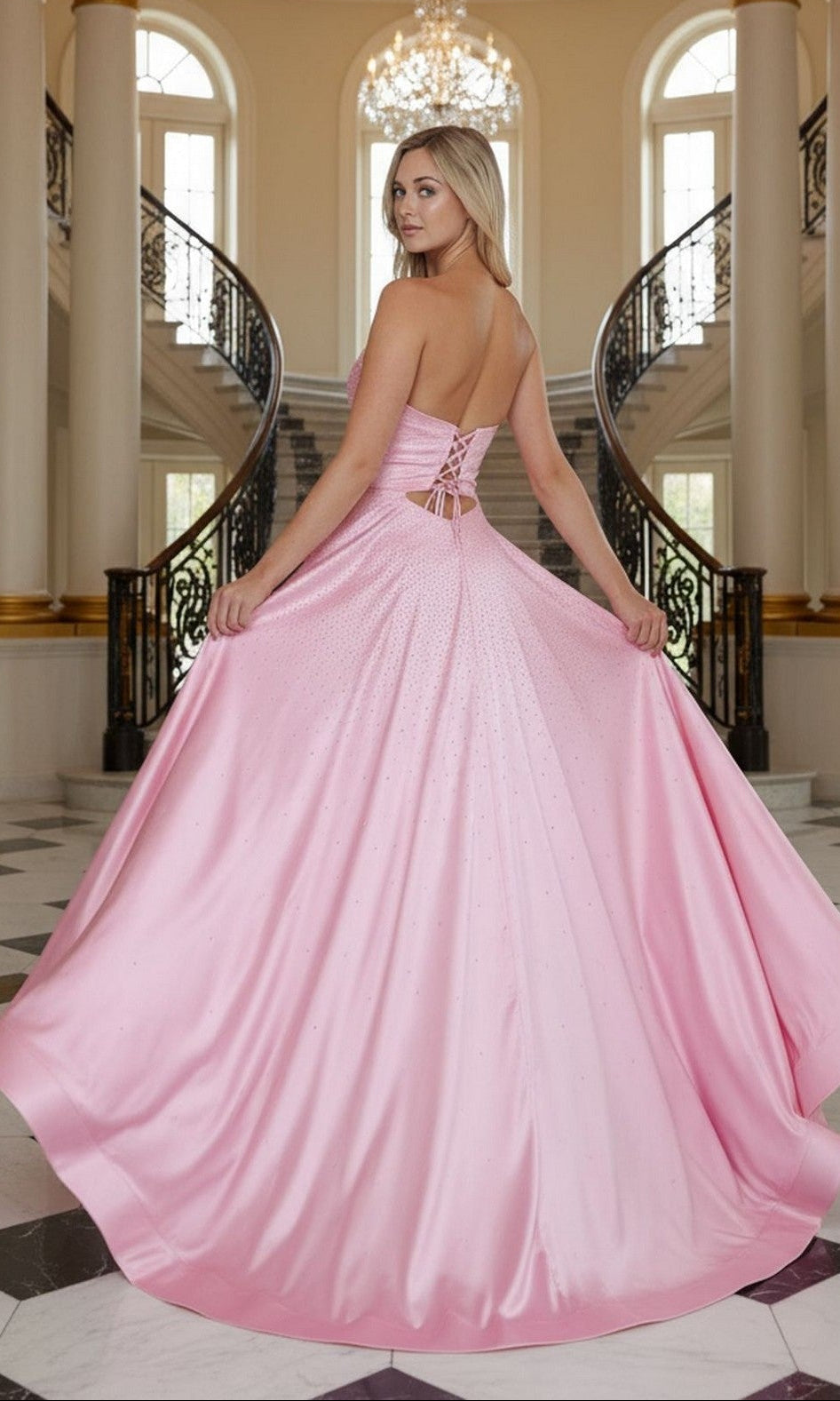 A woman in the Long Prom Dress: Poly USA 25130, a pink strapless satin gown with a sweetheart neckline, stands before a double staircase in an elegant interior with marble floors and tall windows, holding out her dress.
